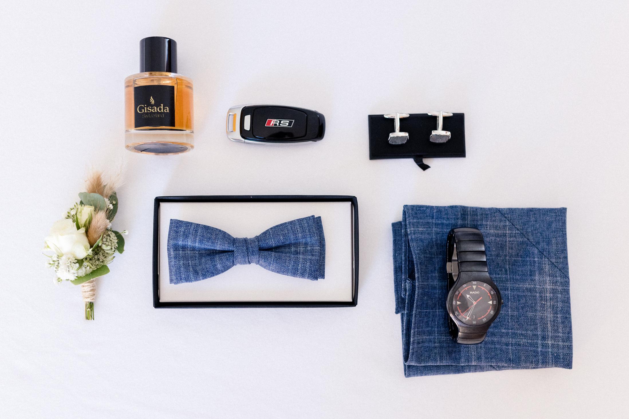Flatlay der Bräutigam-Accessoires – Parfum, Autoschlüssel, Manschettenknöpfe, blaue Fliege, Uhr und Ansteckblume
