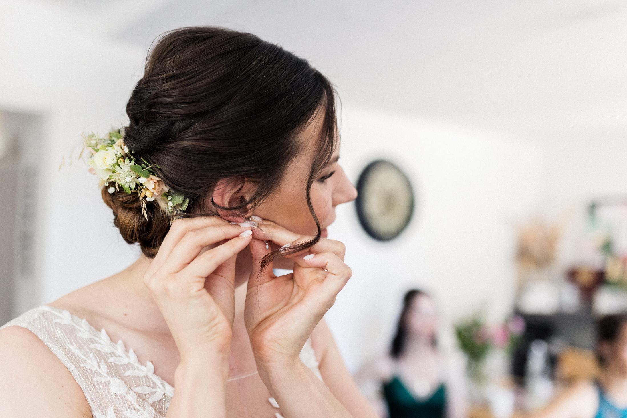 Braut steckt sich die Ohrringe an, Blumenschmuck in der Hochsteckfrisur, Freundinnen im Hintergrund