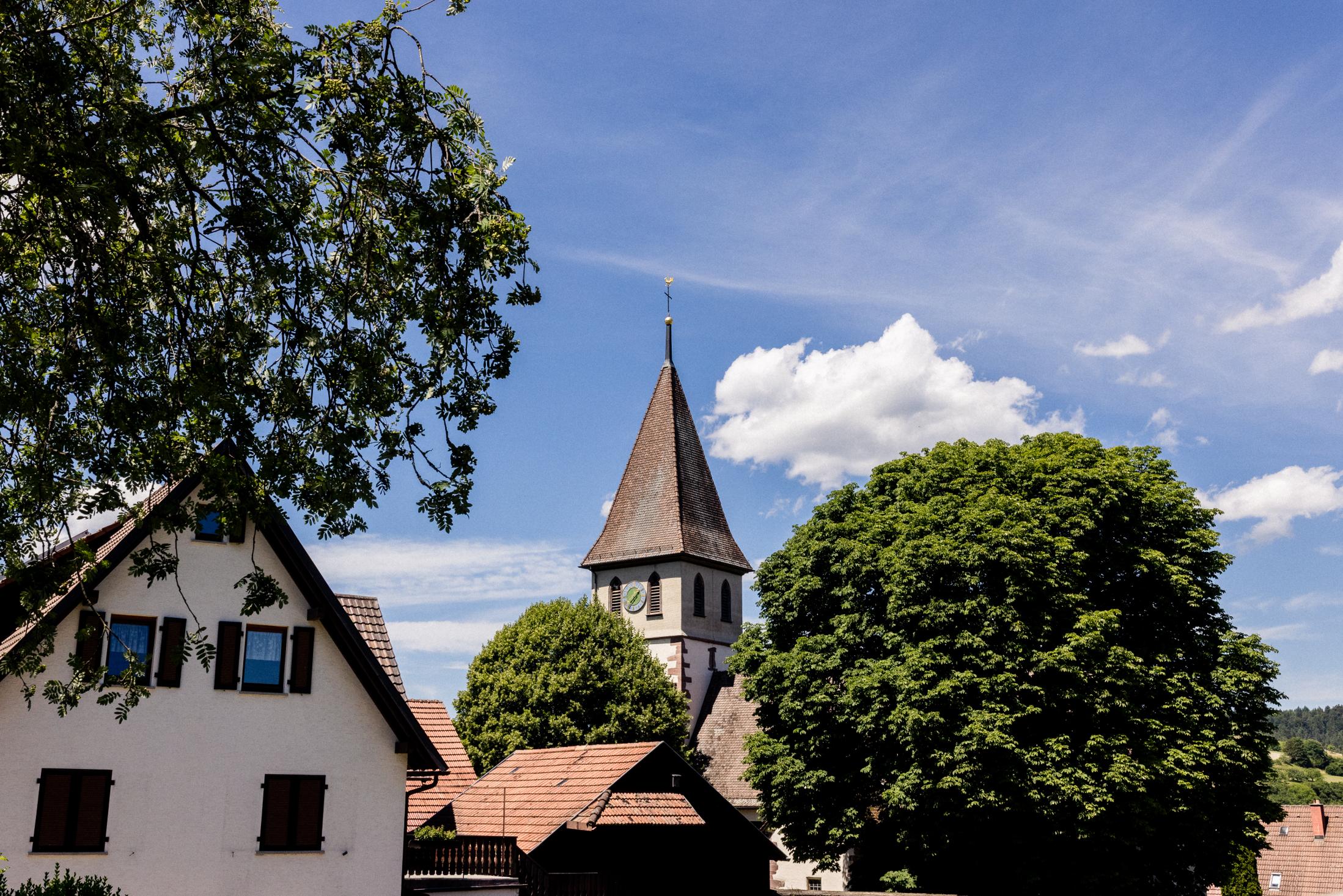 Kirchturm mit spitzem Dach und Uhr hinter Bäumen unter blauem Himmel, Fachwerkhaus im Vordergrund