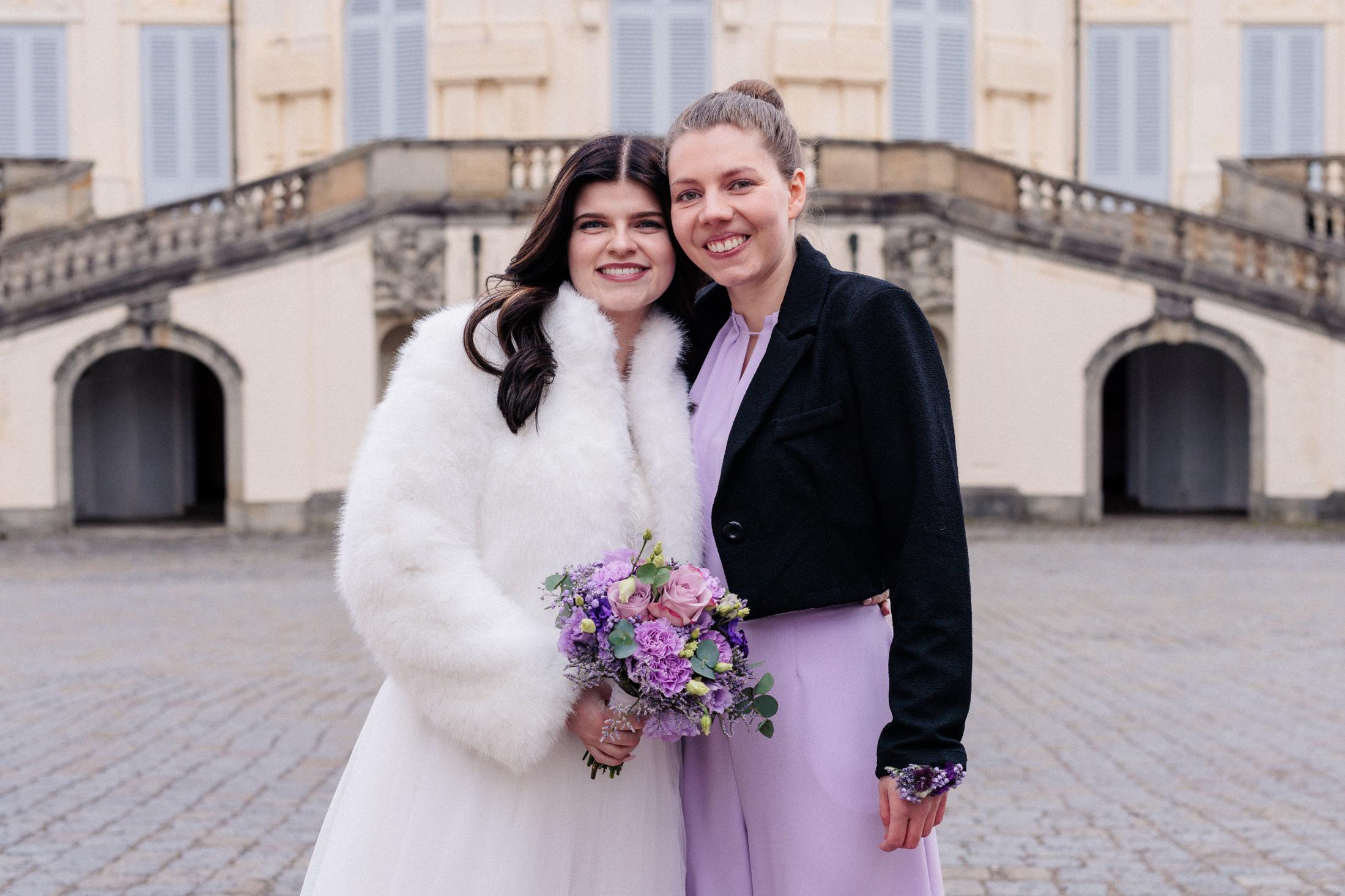 Braut im weißen Fellmantel Wange an Wange mit einer Verwandten, beide lächelnd, Brautstrauß mit lila Rosen im Bild