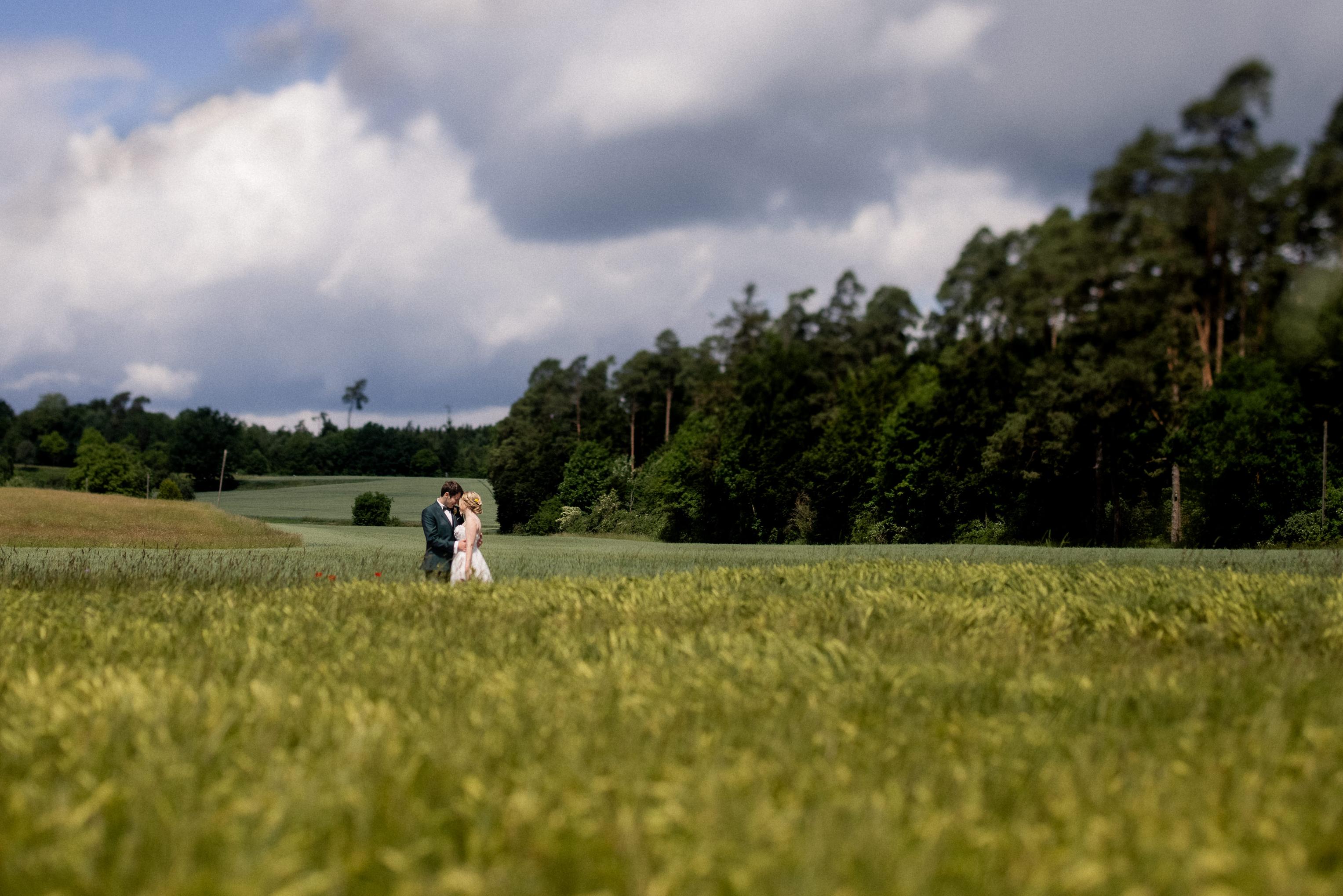 Brautpaar küsst sich in weitem Getreidefeld mit Waldrand und Wolkenhimmel bei Mötzingen