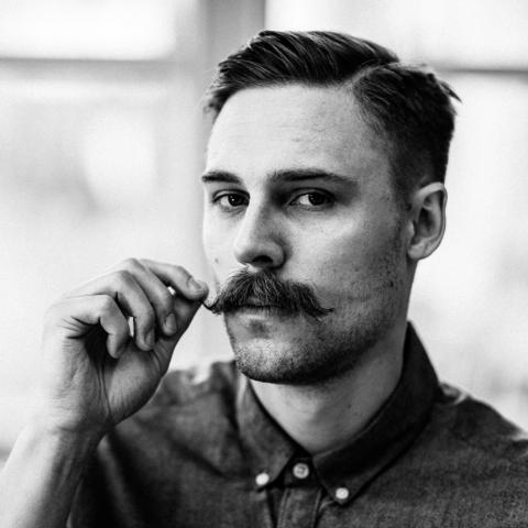 Schwarz-Weiß-Porträt von Daniel Sattler mit Schnurrbart, der nachdenklich in die Kamera blickt.