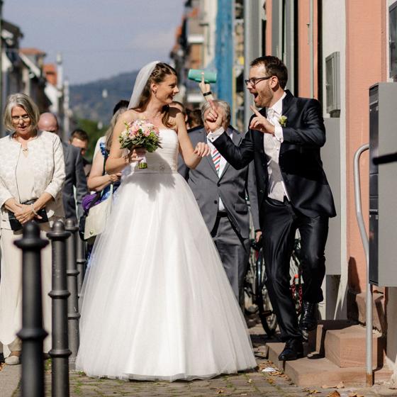 Brautpaar tanzt ausgelassen in einer sonnigen Altstadtgasse, ein Gast hält eine Musikbox über die Köpfe, die Hochzeitsgesellschaft folgt dahinter