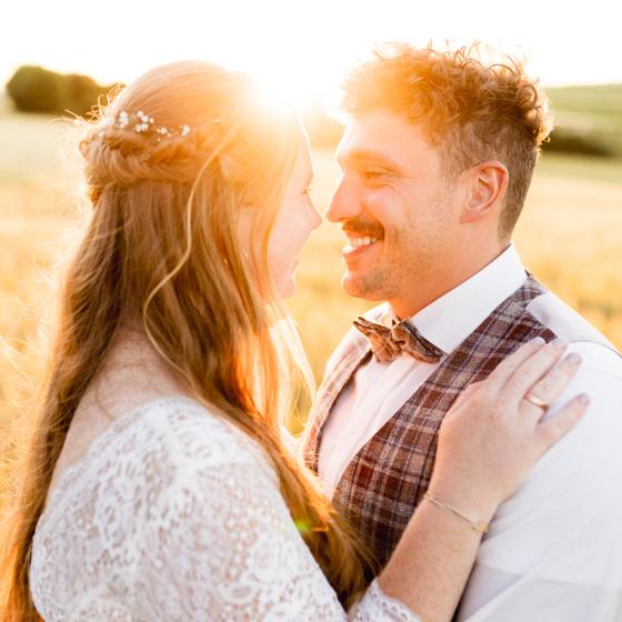 Brautpaar im warmen Gegenlicht kurz vor Sonnenuntergang, eng beieinander auf einer offenen Wiese – Paarfoto in der Golden Hour