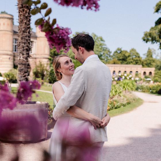 Brautpaar umarmt sich im sommerlichen Schlossgarten zwischen pink blühenden Sträuchern, die Braut schaut lachend zum Bräutigam auf, im Hintergrund Schlossturm und Orangerie