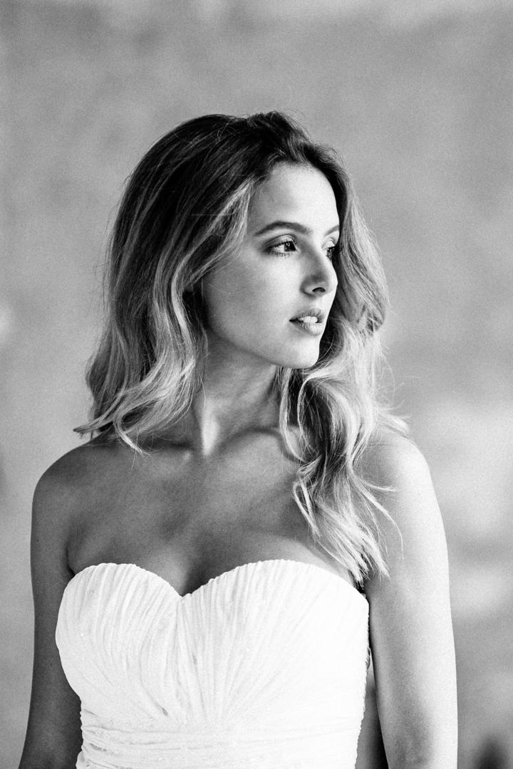 Braut in elegantem, schulterfreiem Brautkleid blickt verträumt in die Ferne, mit weichem Licht und heller, minimalistischer Kulisse.