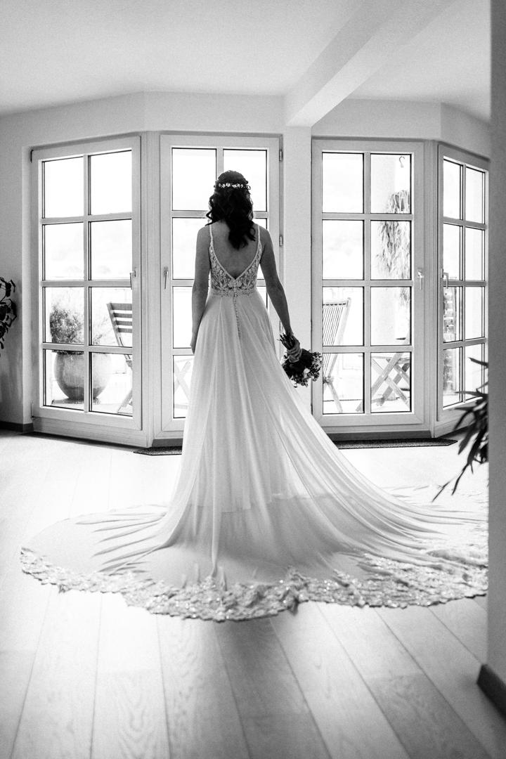 Braut steht in einem eleganten weißen Kleid mit langer Schleppe vor bodentiefen Fenstern und blickt hinaus in die Natur.