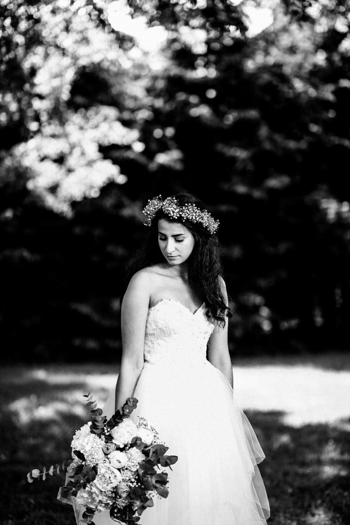 Braut in einem weißen Hochzeitskleid mit Blumenkranz im Haar, posiert mit einem eleganten Brautstrauß in einer sonnigen Gartenkulisse.