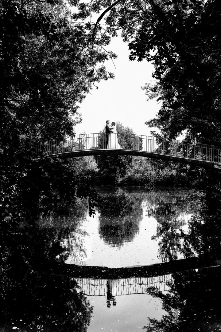 Brautpaar steht romantisch auf einer Brücke über einem ruhigen Teich, eingerahmt von üppigem Grün, mit Spiegelung im Wasser.