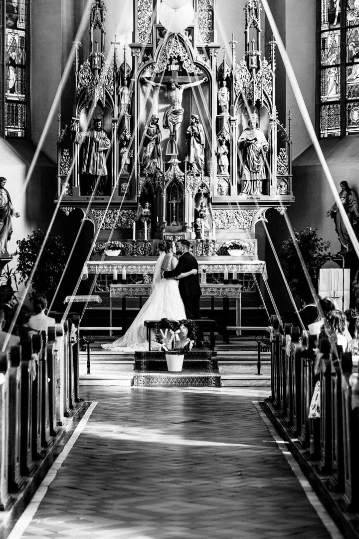 Brautpaar steht vor einem reich verzierten Altar mit leuchtenden Farben und Figuren in einer Kirche, während der Zeremonie.