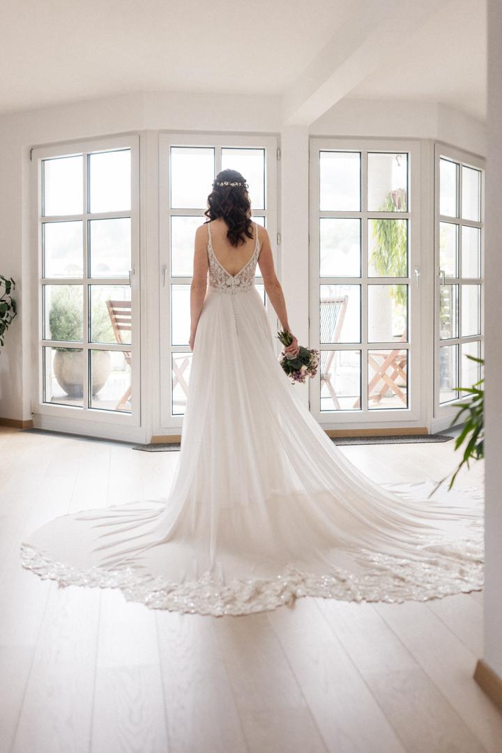 Braut steht in einem eleganten weißen Kleid mit langer Schleppe vor bodentiefen Fenstern und blickt hinaus in die Natur.