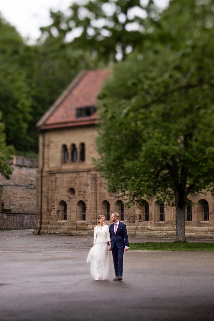 Brautpaar spaziert Hand in Hand vor einem historischen Gebäude, umgeben von Bäumen in einer ruhigen Kulisse.