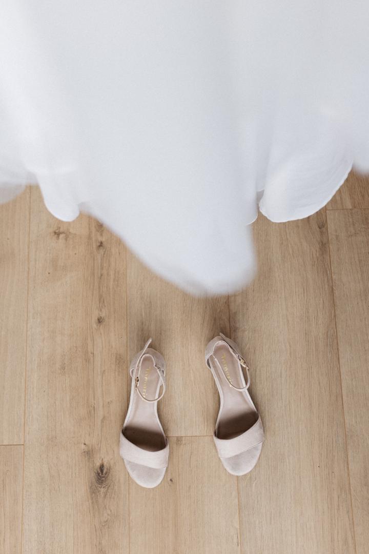 Nude-farbene Brautschuhe mit Riemchen auf hellem Holzboden, darüber der Saum des Brautkleids