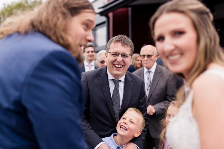 Braut und Bräutigam lachen mit ihren Gästen, während ein kleiner Junge freudig im Vordergrund steht.