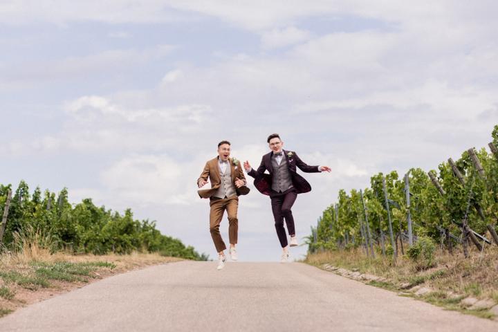 Zwei Bräutigame beim Paarshooting in den Weinbergen, beide springen synchron in die Luft und lachen fröhlich.