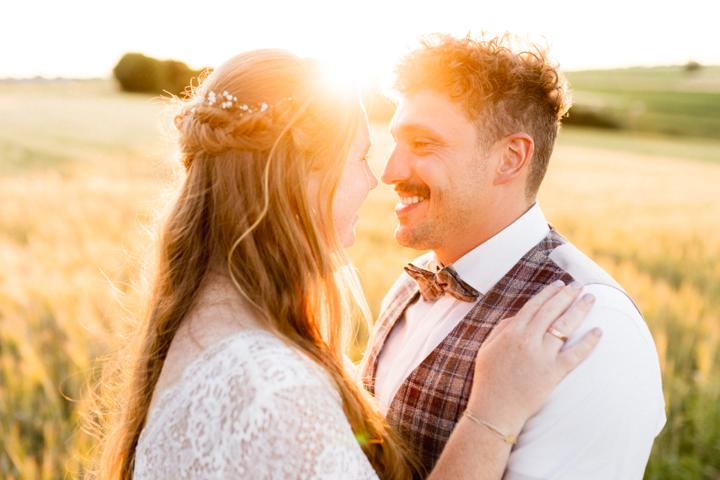 Brautpaarshooting beim Sonnenuntergang: Braut im Spitzenkleid mit locker geflochtenem Haar und Bräutigam in kariertem Gilet und Fliege stehen in einem goldenen Weizenfeld bei untergehender Sonne und lächeln sich verliebt an.