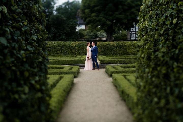 Brautpaar steht romantisch in einem gepflegten Barockgarten mit symmetrischen Hecken und einem historischen Fachwerkhaus im Hintergrund.