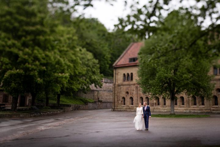 Brautpaar spaziert Hand in Hand auf einer von Bäumen gesäumten Allee vor einem historischen Gebäude.