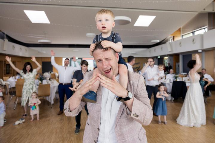 Mann trägt ein Kind auf den Schultern, beide im Mittelpunkt einer fröhlichen Tanzrunde in heller Festhalle, im Hintergrund jubeln Gäste und tanzen mit erhobenen Armen.