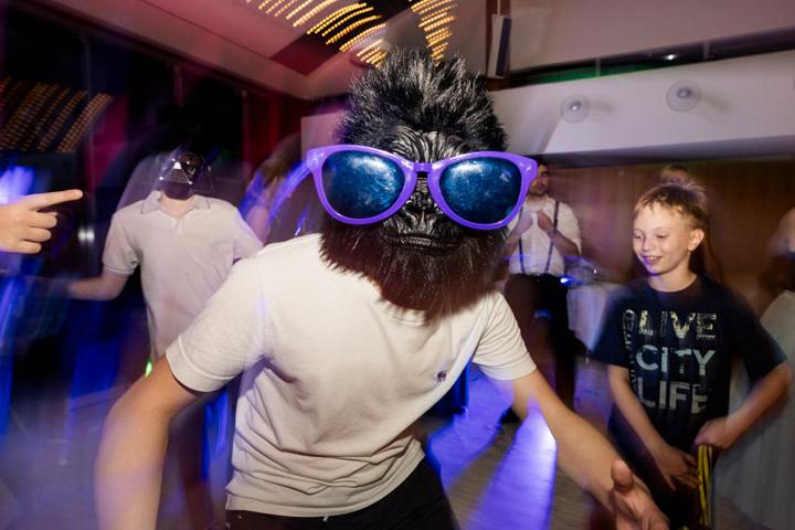 Kind mit großer, verspiegelter Sonnenbrille-Maske tanzt ausgelassen zwischen Erwachsenen, wird von verblassten Lichterprojektionen umspielt und sorgt für heitere Überraschung.