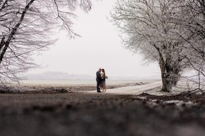 Brautpaar umarmt sich auf einer frostigen Allee, umgeben von schneebedeckten Bäumen und einer winterlichen Landschaft.