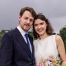 Brautpaar-Shooting beim Riverhouse in Stuttgart – der Bräutigam im dunkelblauen Anzug mit blauer Krawatte und die Braut im schlichten weißen Kleid mit Blumenkranz lächeln eng umschlungen in die Kamera, im Vordergrund ein bunter Brautstrauß.